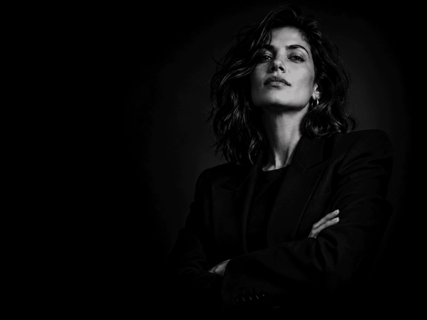 Black and white portrait of a woman in a black blazer, arms crossed, lit against a dark background